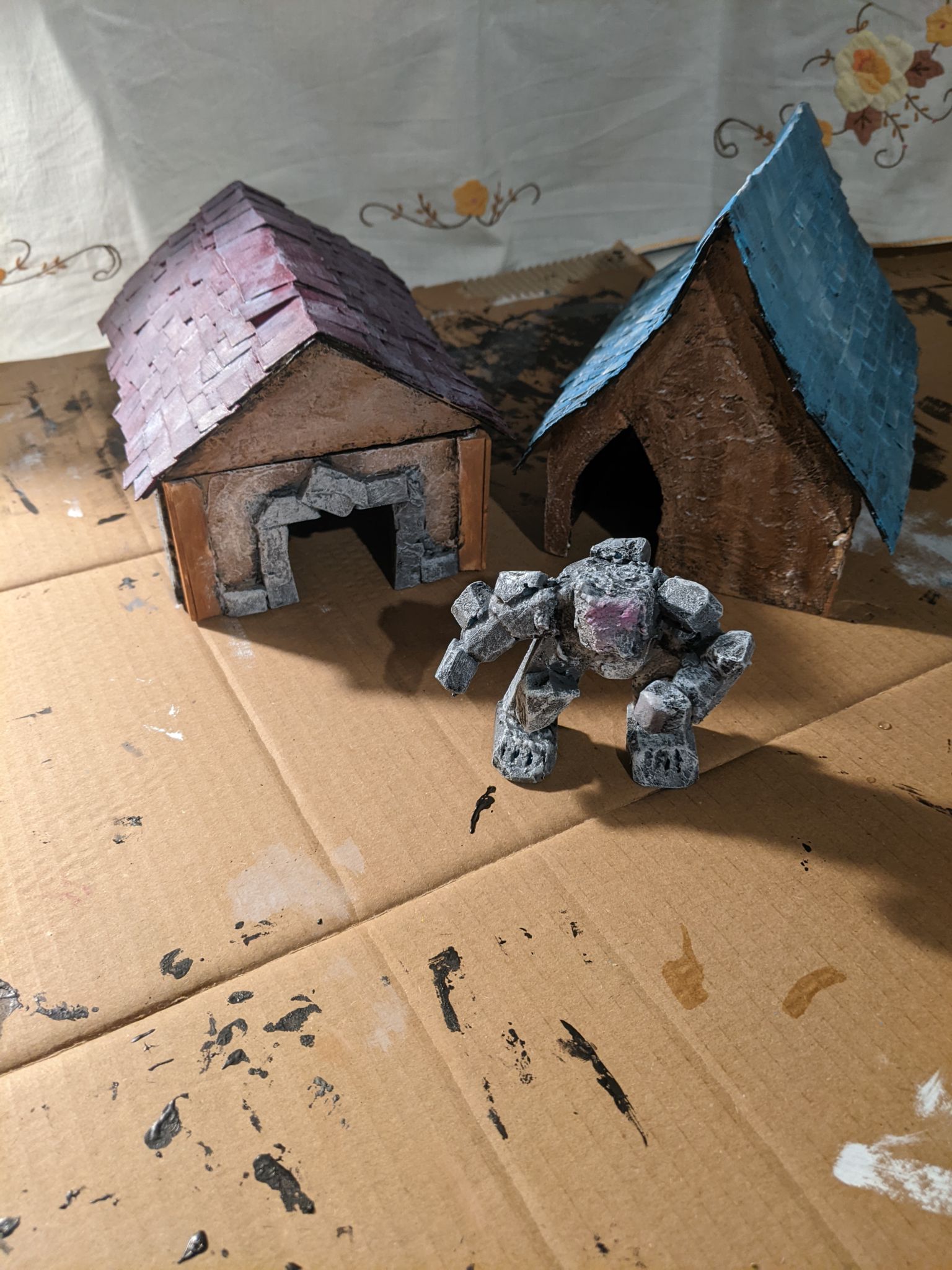 A picture of two painted miniature houses for a TTRPG. In front of them is a self made stone golem consisting of painted XPS foam.
