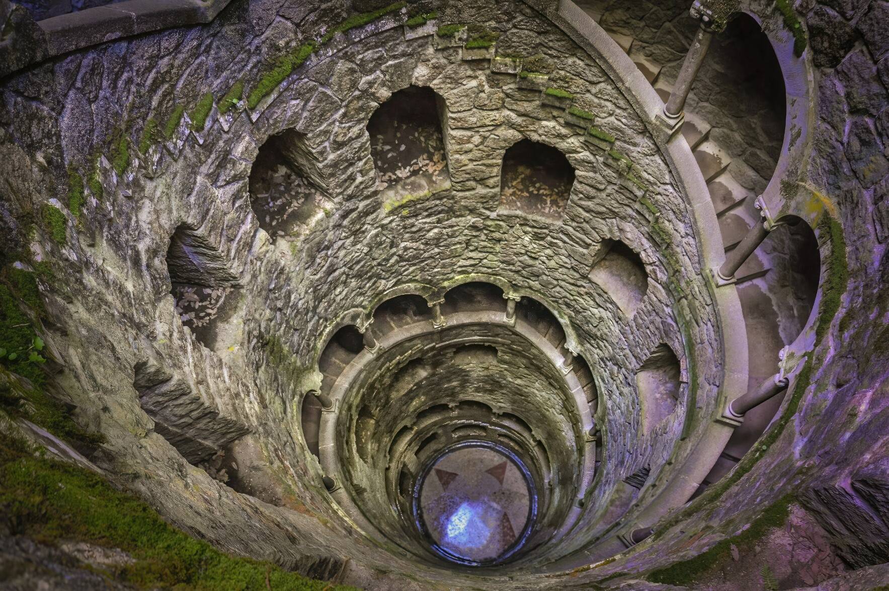 Deep spiral staircase into the Initiation Well. Built of stone, covered in green moss lead down many floors to tile floor at the bottom of the well.